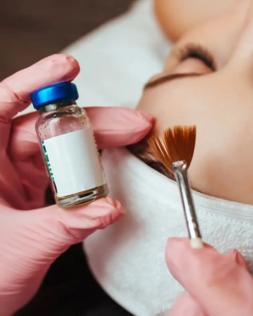 Chemical Peel Recovery: How Long Does It Take? 11 Aesthetician in pink gloves holding a medical-grade chemical peel recovery vial and fan brush near a client's face during a skin resurfacing treatment at Two Chicks Aesthetics.